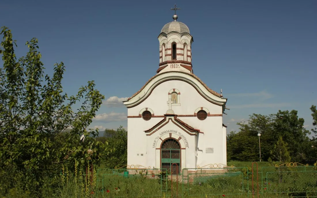 Бял селски храм с червен купол и каменен градеж, увенчан с кръст, сред зеленина в Калояновско