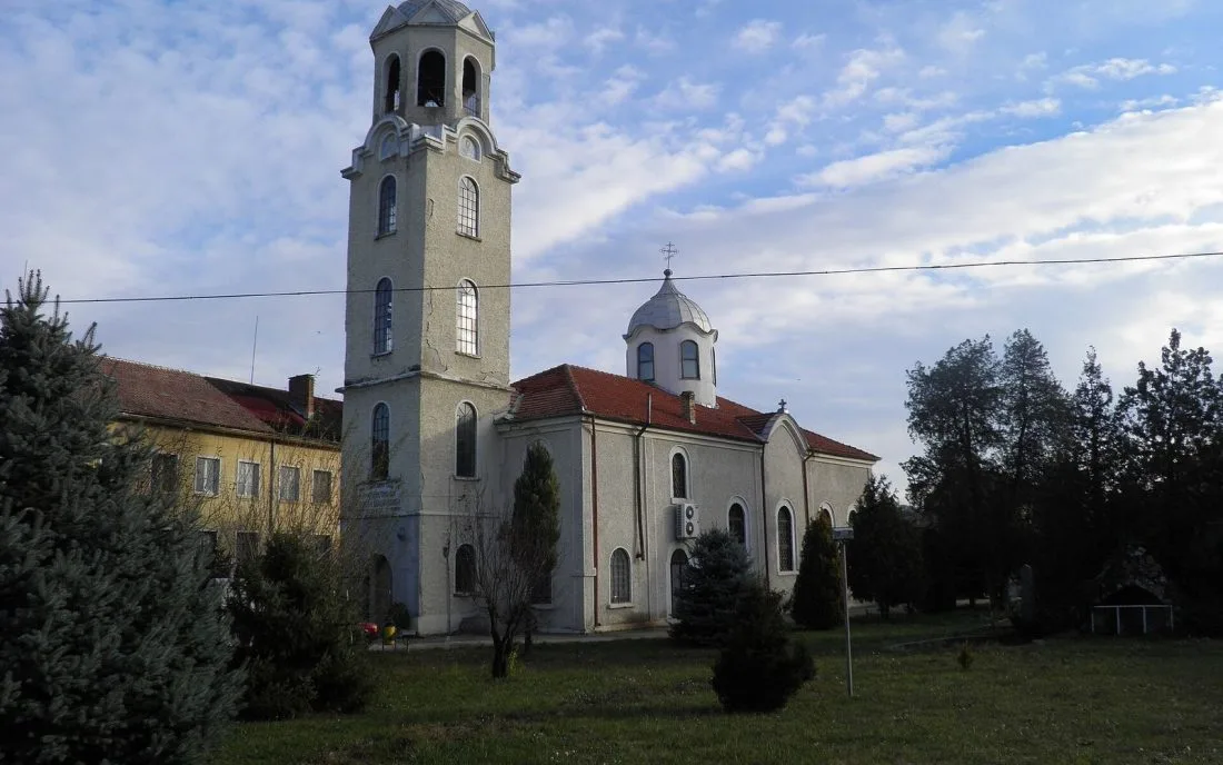 Голям бял православен храм с висока камбанария, купол и червен покрив, сред иглолистни дървета