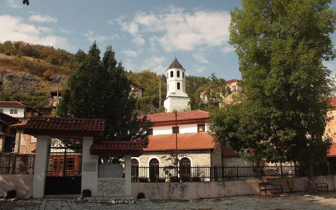 Църквата Свети Георги в Беловския край с бялата камбанария, керемидените покриви и каменния двор под скалист склон