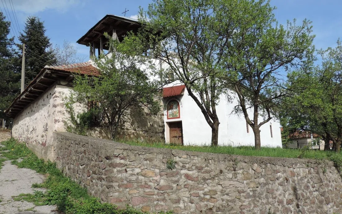 Стара каменна православна църква с дървена камбанария и икона на конник над входа, зад нисък каменен зид