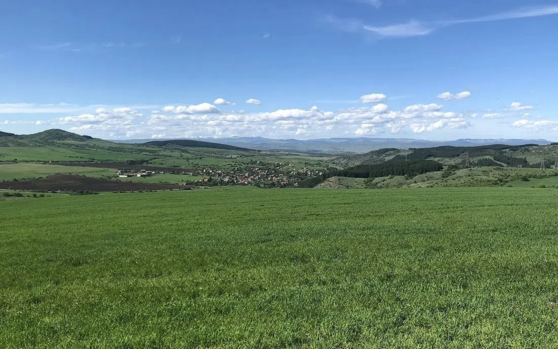Широка зелена поляна край Божурище със село в далечината и планини на хоризонта