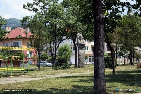 Малък парк с паметник в Бухово, зад него къщи и залесени старопланински склонове